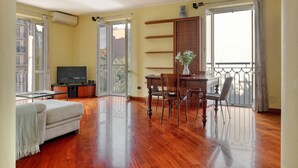 A spacious living room with hardwood floors, a sofa, a dining table with chairs, a TV, and a balcony with a view of buildings. - Delightful 2BR flat near Navigli (Milan)
