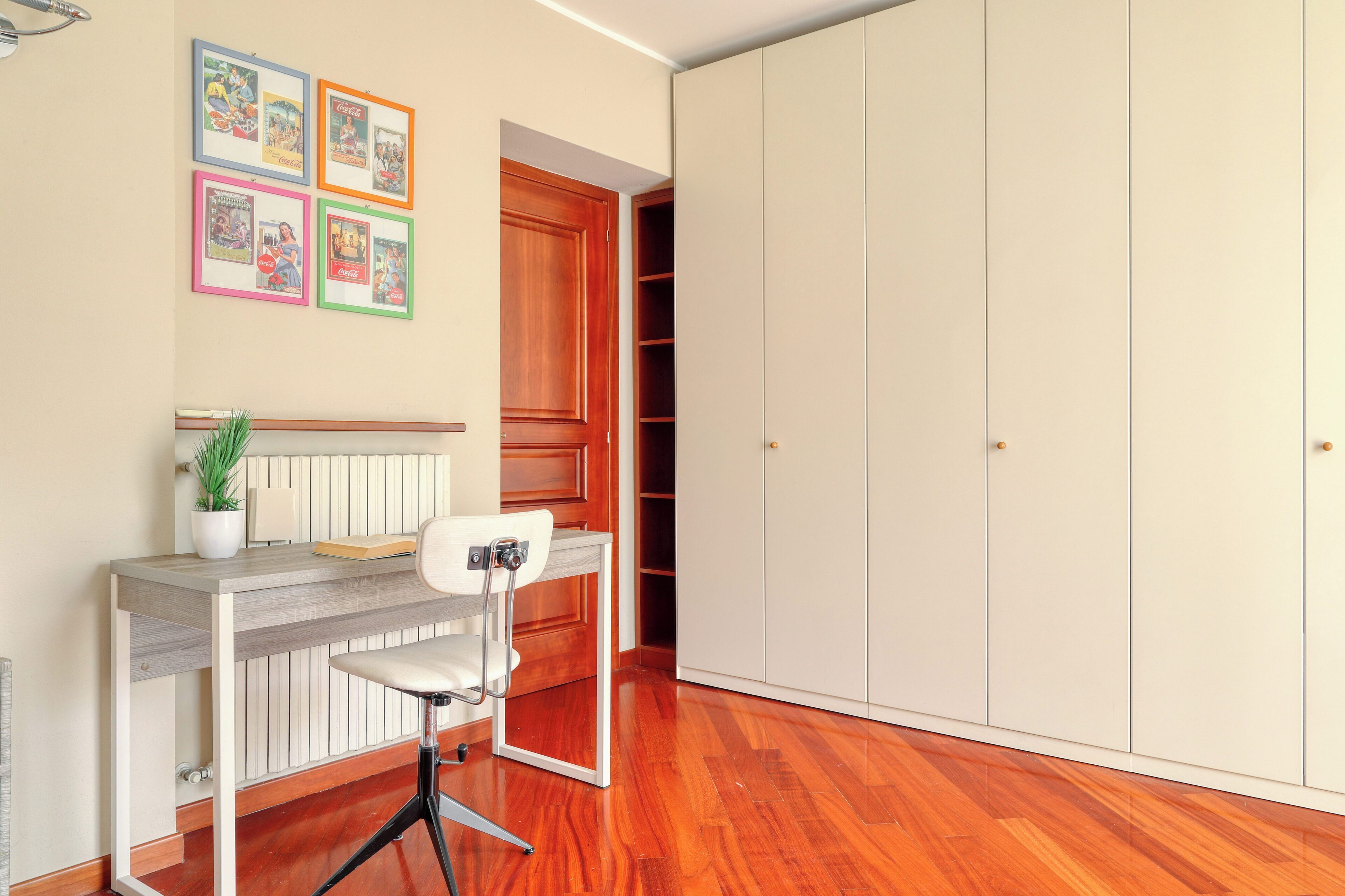 Ein Zimmer mit Holzboden, Schreibtisch mit Stuhl, Blumentopf und Kleiderschrank.