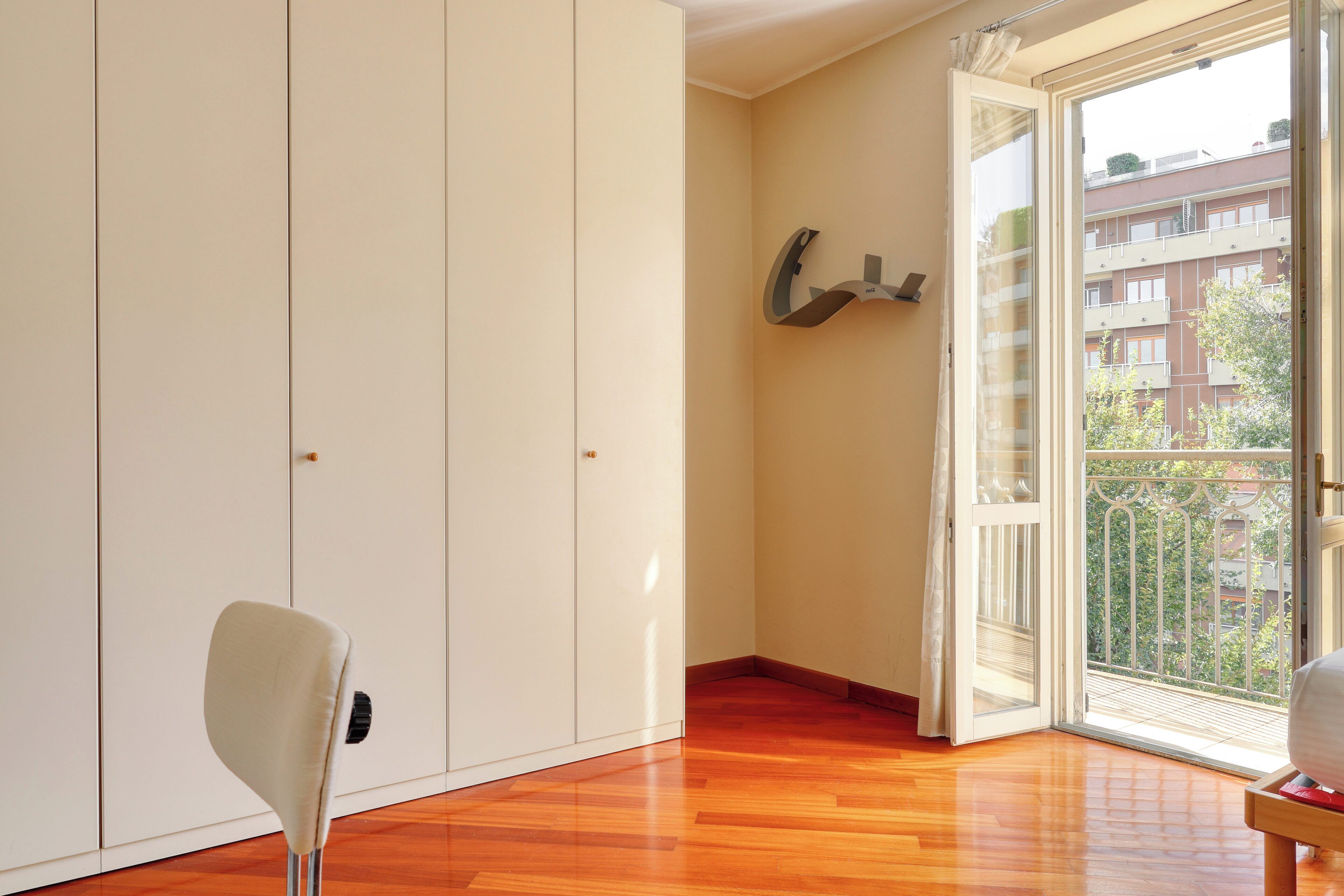 Een kamer met een houten vloer, een witte kast en een schuifglazen deur die uitkomt op een balkon met uitzicht op gebouwen.