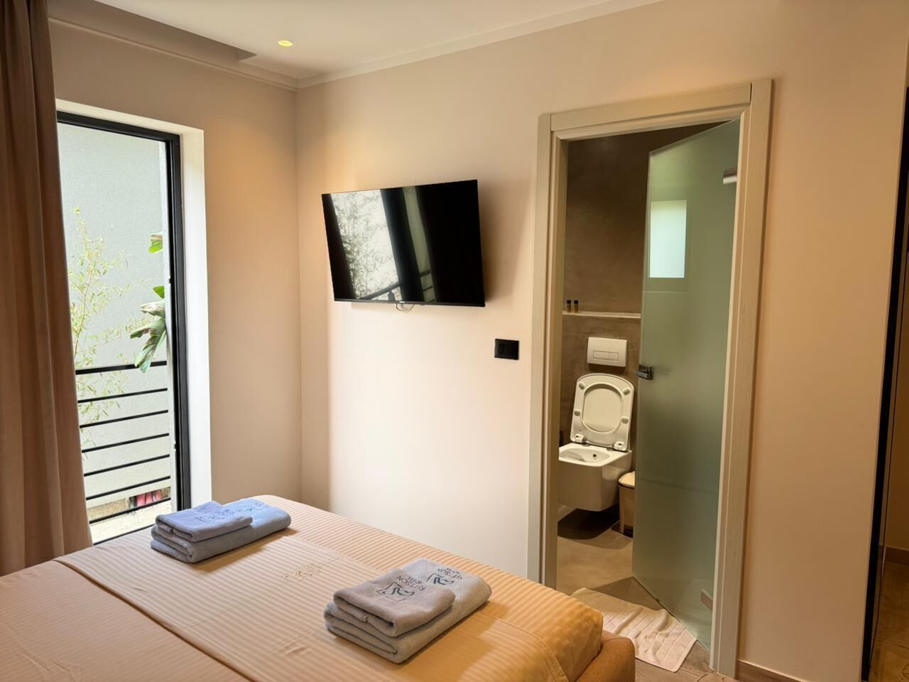 A hotel room with a bed, a TV mounted on the wall, a bathroom with a toilet and sink, and a sliding glass door to the outside.