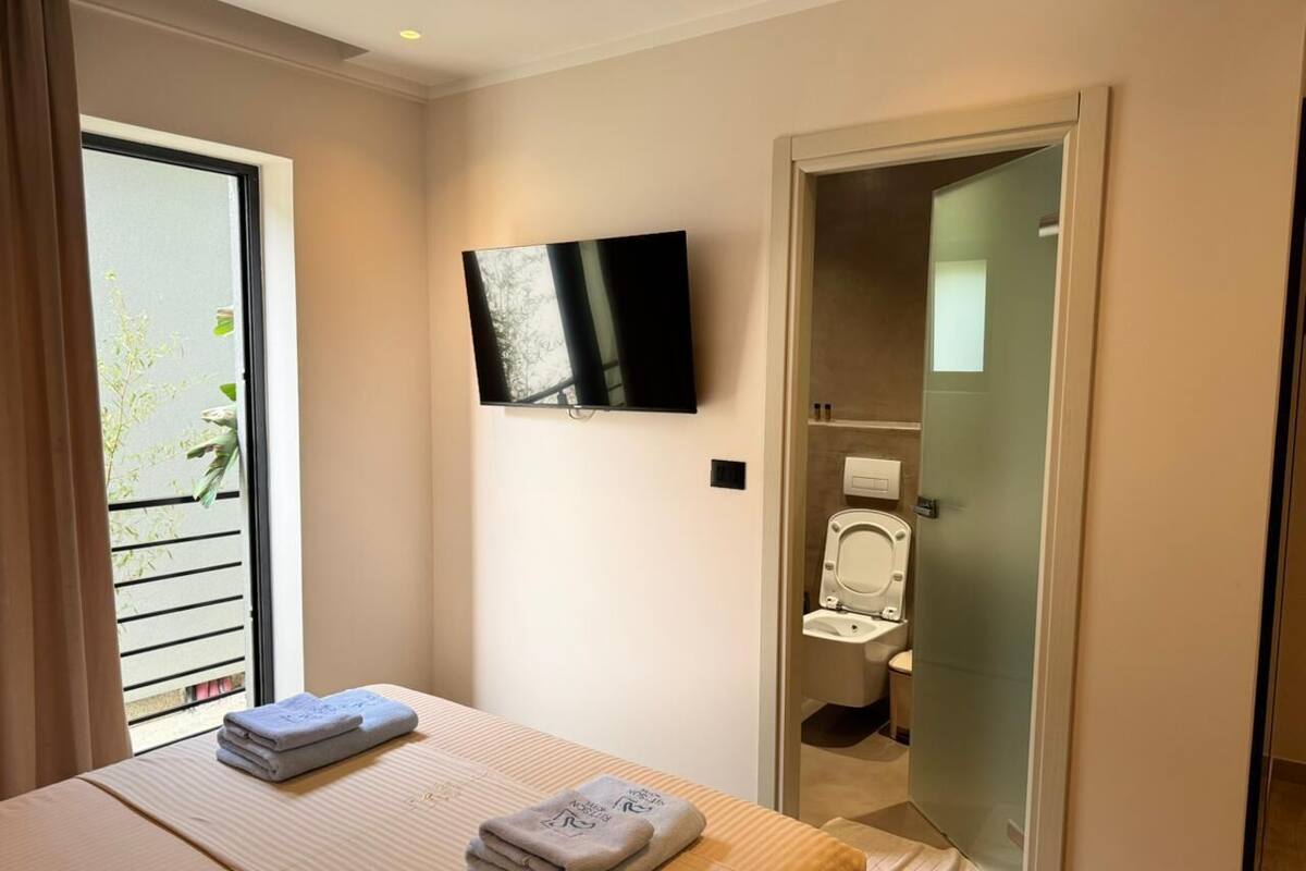 A hotel room with a bed, a TV mounted on the wall, a bathroom with a toilet and sink, and a sliding glass door to the outside.