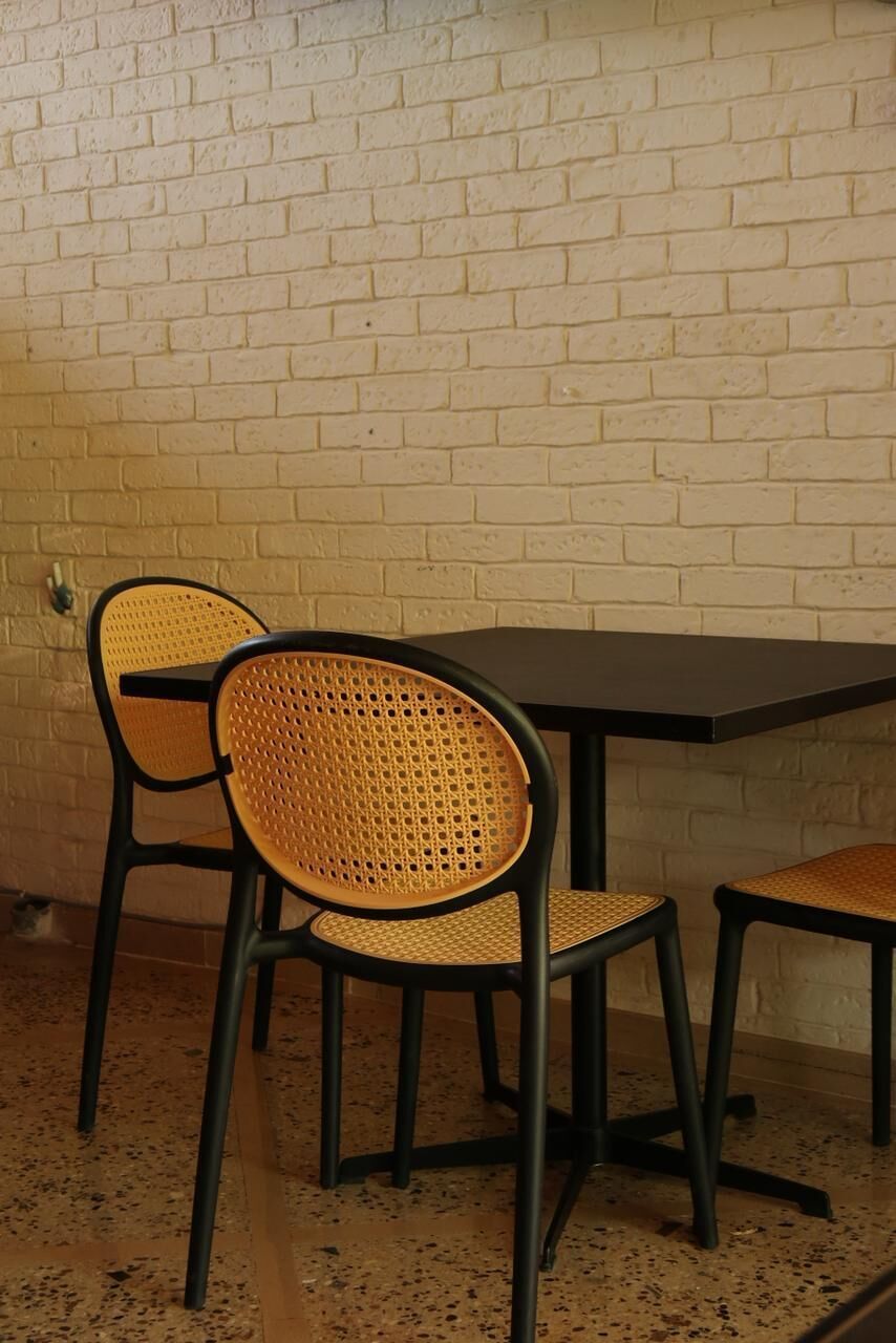 Een tafel en twee stoelen met gevlochten zittingen, een zwart tafelonderstel en een witte bakstenen muur.