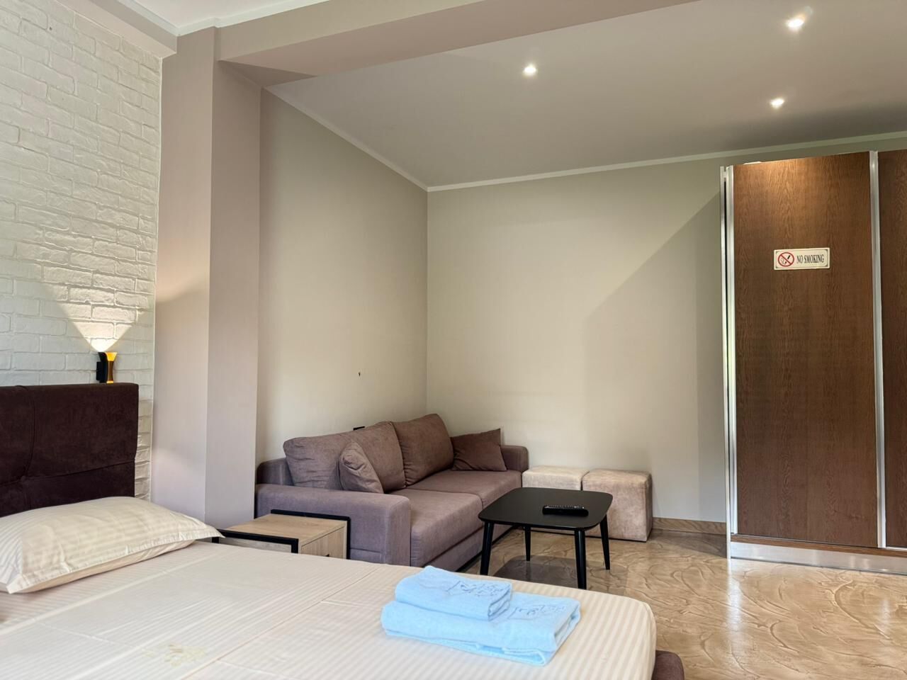 A hotel room with a bed, a sofa, a small table, a lamp, and a sliding door with a 'No Smoking' sign.