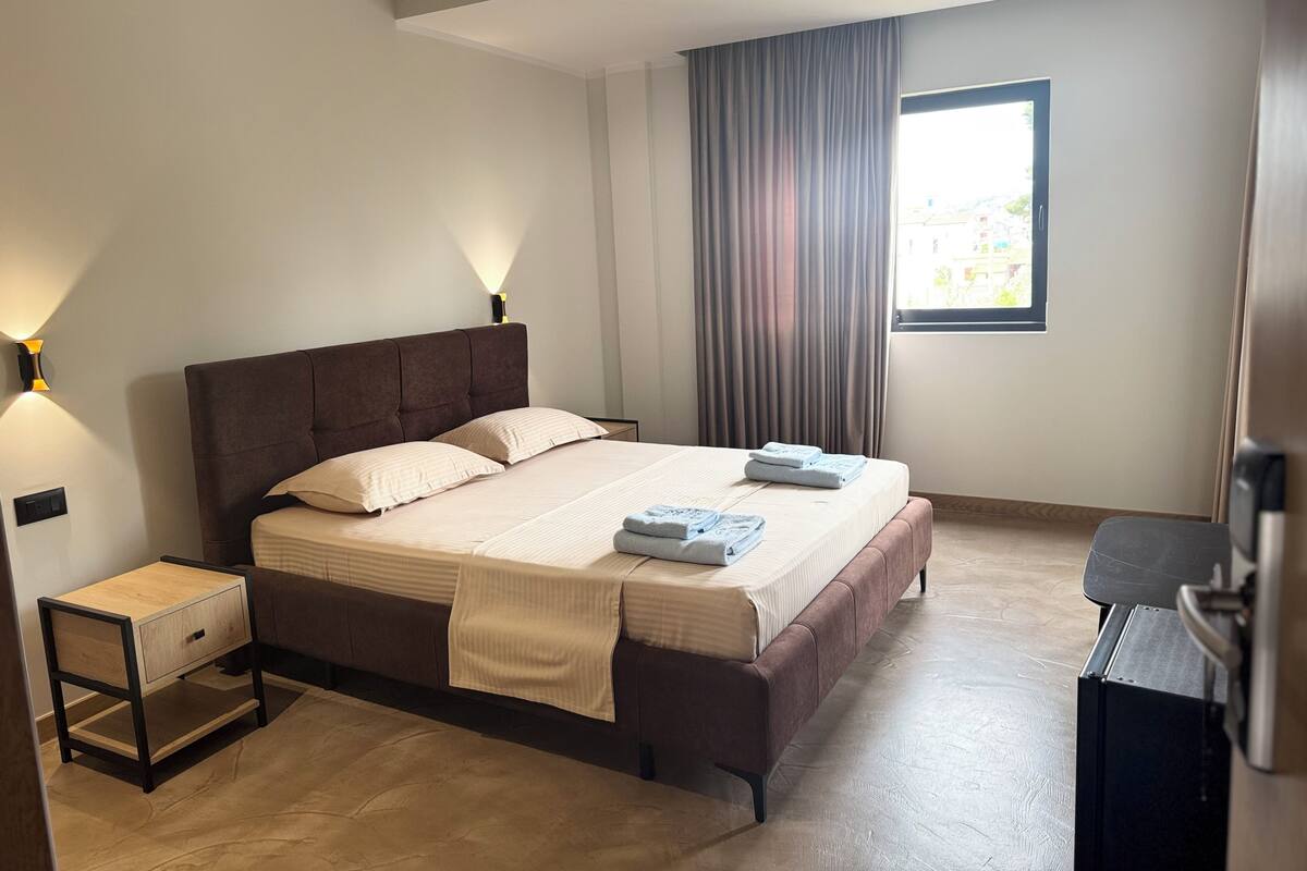 A neatly arranged bedroom with a bed, bedside tables, a desk, and a window with curtains.