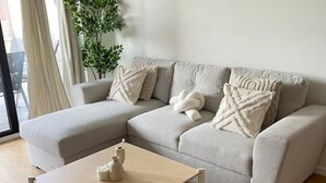 Ein modernes Wohnzimmer mit einem hellgrauen Ecksofa, zwei weißen Kissen, einem kleinen Holztisch mit zwei weißen Vasen und einer Topfpflanze.