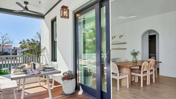 A dining area with a wooden table and chairs, a potted plant, and a sliding glass door leading to a balcony.