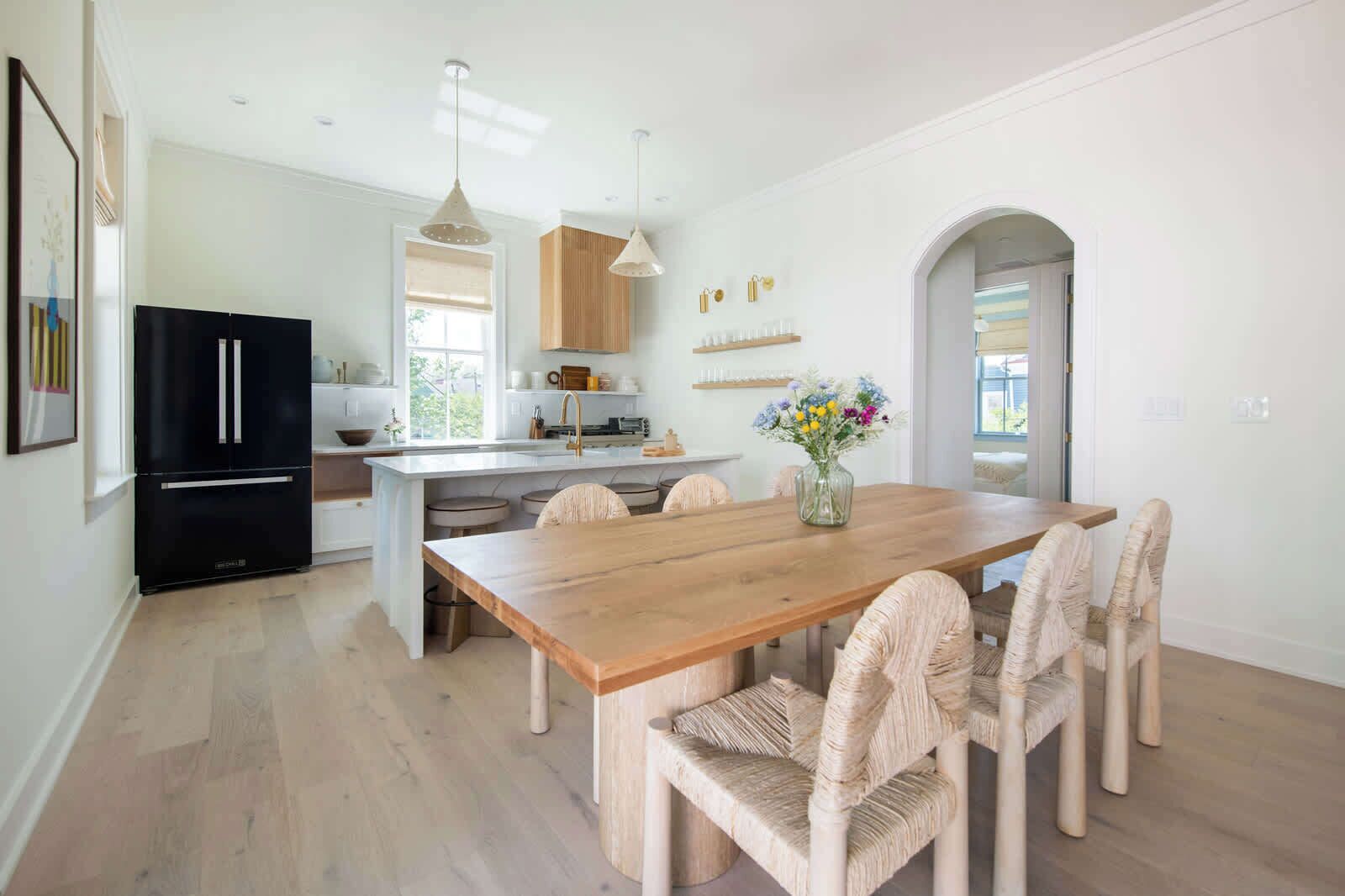 A modern kitchen with a central island, black refrigerator, and wooden dining table with wicker chairs.