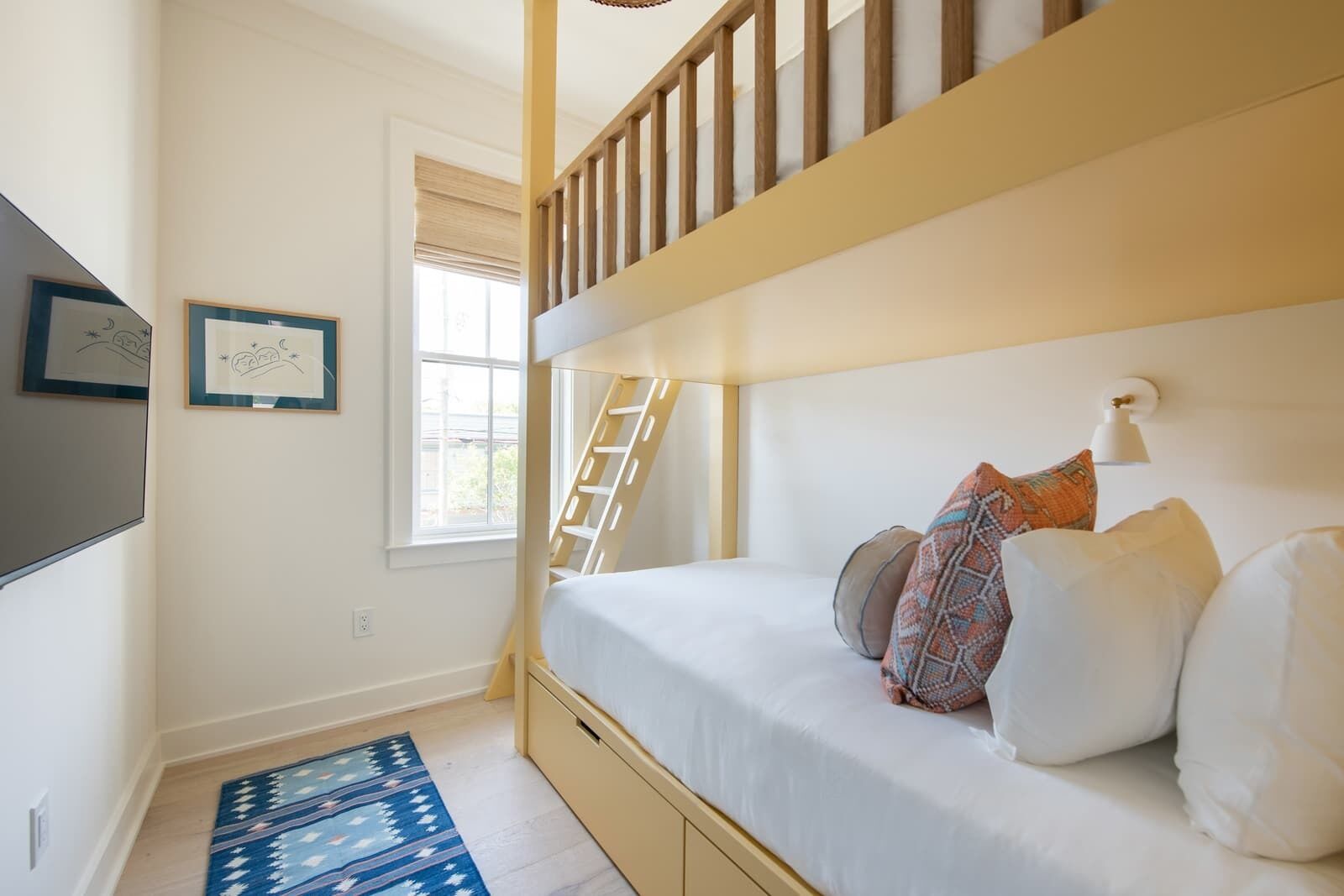 A bunk bed room with a ladder, a window with blinds, a framed picture, and a patterned rug.