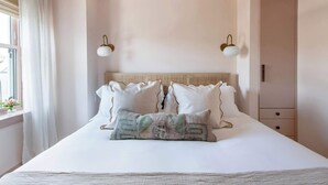 Un lit bien fait, avec une literie blanche et des coussins décoratifs, une tête de lit à motifs tissés et une armoire de couleur claire sur le côté droit.