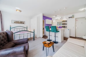 A modern room with a bed, a leather armchair, a small wooden table, and a kitchen area with a purple wall and white cabinets.