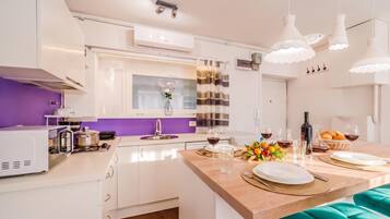 A modern kitchen with white cabinetry, a purple accent wall, and a dining area with a teal bench.