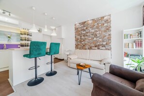 A modern living room with a white sofa, two green bar stools, a wooden coffee table, and a brown armchair. A stone wall and bookshelf with books are visible in the background. - Charming Downtown Studio (Zagreb)