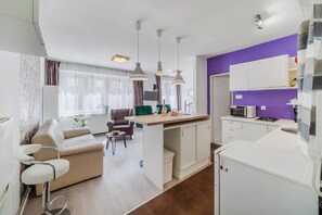 A modern kitchen with white cabinets, a purple accent wall, and a dining area with a sofa and chairs.
