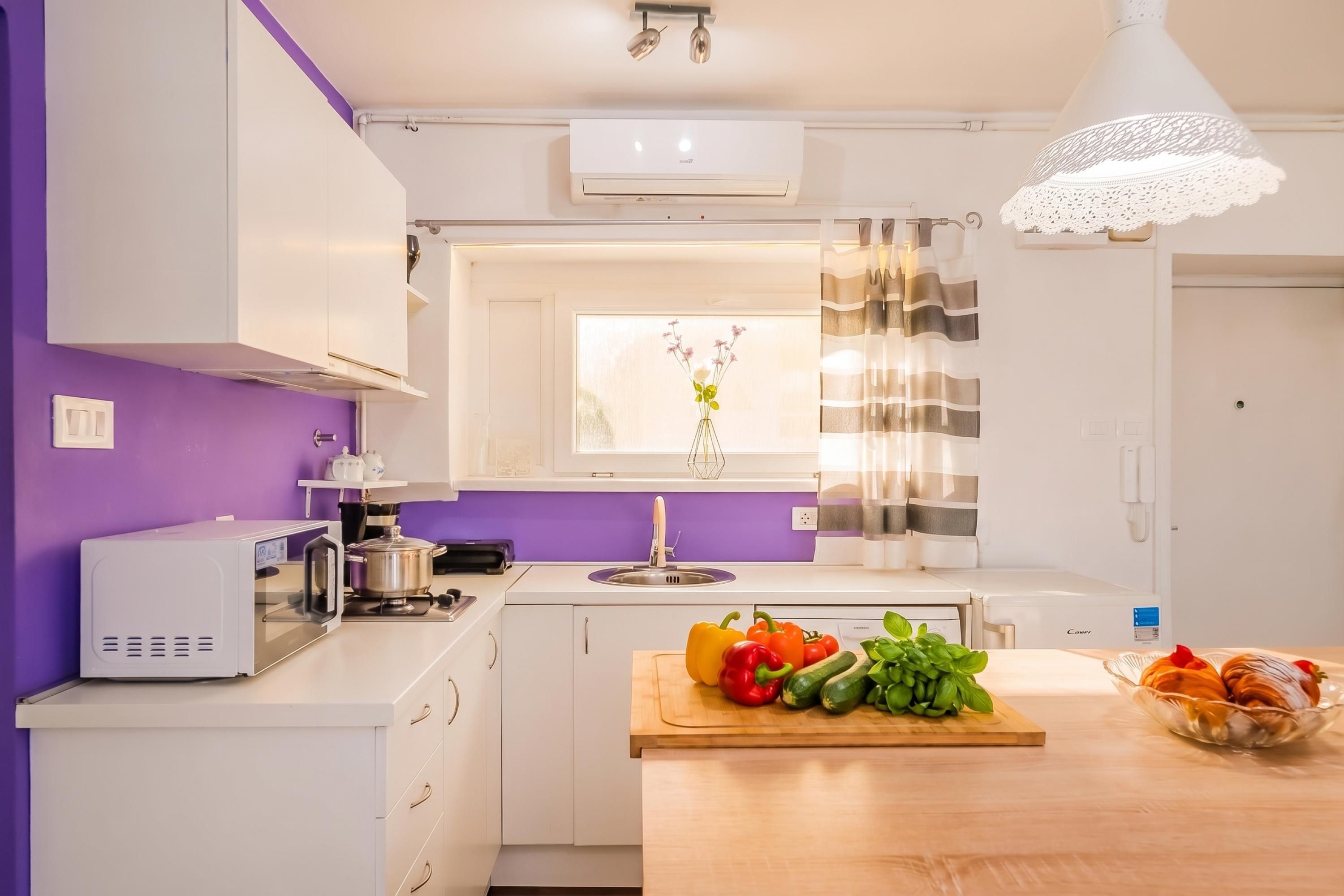 A modern kitchen with white cabinets, a purple wall, a window with a view, and a wooden countertop with vegetables.