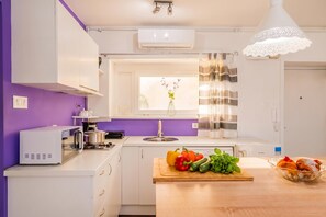 A modern kitchen with white cabinets, a purple wall, a window with a view, and a wooden countertop with vegetables. - Charming Downtown Studio (Zagreb)