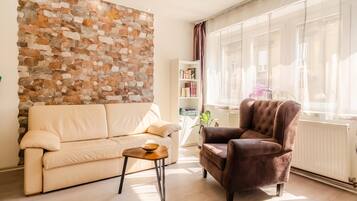 A living room with a beige sofa, a brown armchair, a small table with a bowl, a bookshelf, and a stone wall.