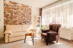 A living room with a beige sofa, a brown armchair, a small table with a bowl, a bookshelf, and a stone wall. - Charming Downtown Studio (Zagreb)