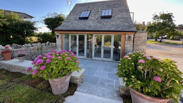 A small stone house with a sloped roof, large windows, and a paved patio area with potted plants. - Idyllic secluded hideaway with countryside views (Hailey)