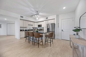 A modern kitchen with white cabinets, a stainless steel refrigerator, a central island with bar stools, and a ceiling light fixture. - Luxury Condo with Sanctuary Views Merritt Island (Merritt Island)