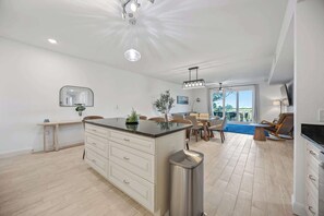 A modern kitchen with a central island, a dining area with chairs, a small table, and a view through the glass door. - Luxury Condo with Sanctuary Views Merritt Island (Merritt Island)