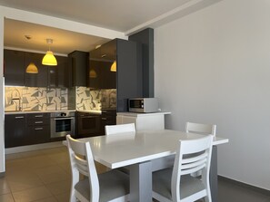 Une cuisine moderne avec des meubles de couleur sombre, une table à manger blanche et deux chaises. On trouve un four à micro-ondes sur le plan de travail et un luminaire suspendu au-dessus.