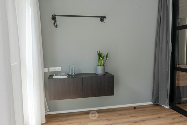 Une pièce moderne avec un mur gris, un plancher en bois, une étagère murale en bois foncé sur laquelle est posée une plante en pot, une petite table avec un livre et des bouteilles, et une tringle à rideaux noire.