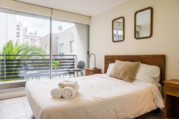 A hotel room with a large bed, two bedside mirrors, a wooden headboard, and a view of a balcony with a chair and plants.