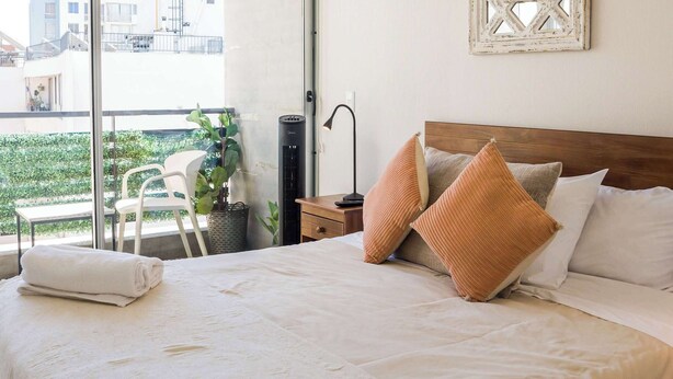 A bedroom with a large bed, a bedside table, a lamp, a chair, and a view of the outside. - Central Chic 1BR Steps from Metro & Attractions (Santiago)
