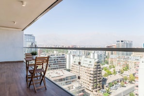 A balcony with a wooden table and chairs, a potted plant, and a cityscape view with mountains in the background. - Chic 2BR Haven in the Heart of Las Condes Oasis (Unknown)