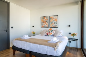 A neatly made bed with white linens and a beige throw, a small bedside table with a vase of flowers, and two framed floral artworks on the wall. - Chic 2BR Haven in the Heart of Las Condes Oasis (Unknown)