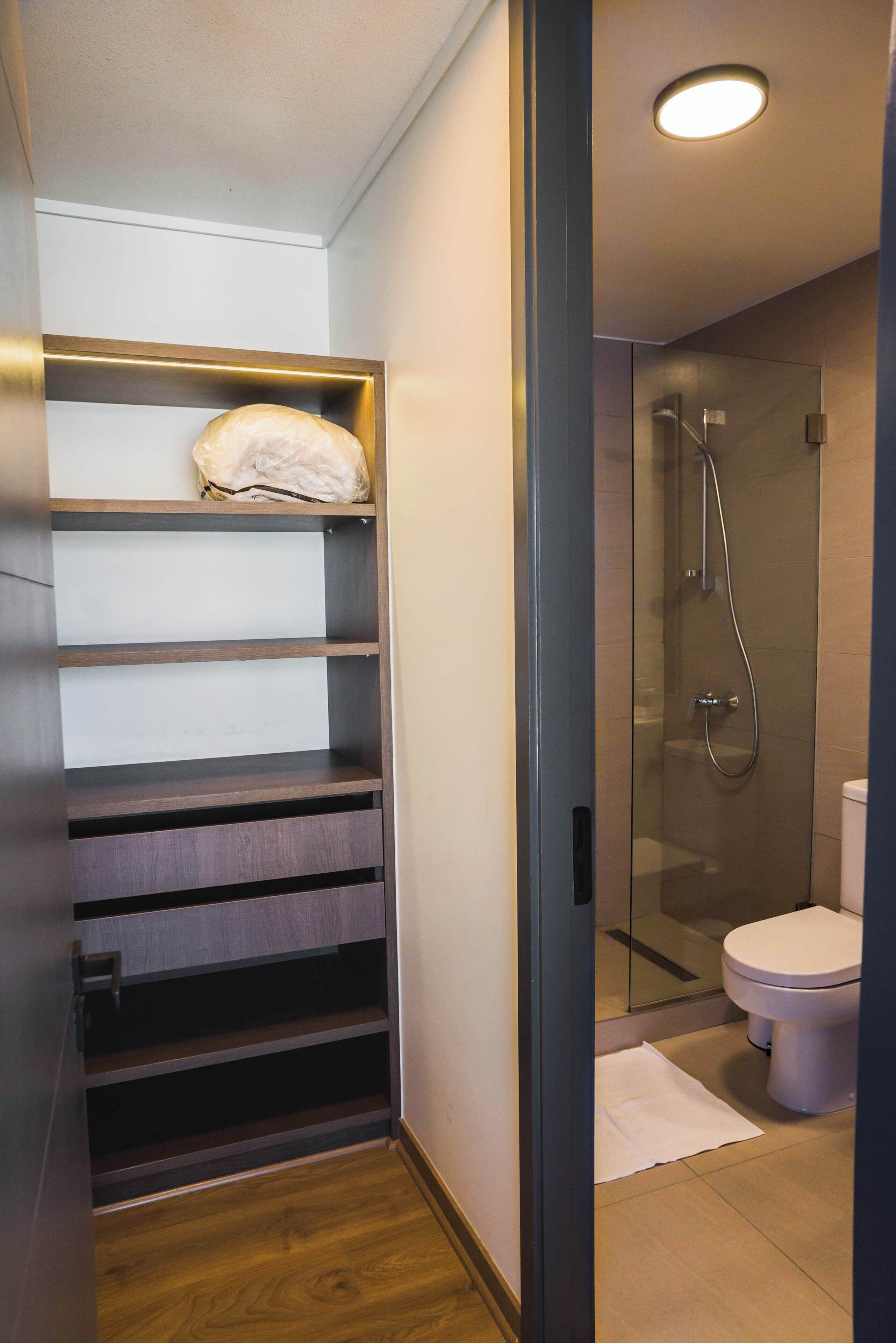 Une salle de bain moderne dotée d’une douche à parois de verre, de toilettes et d’une étagère intégrée en bois où est posée une serviette.
