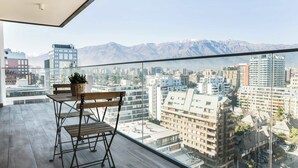 A balcony with a wooden chair and table, a potted plant, and a cityscape view with mountains in the background. - Prime 2BR Oasis in the Heart of Las Condes (Unknown)