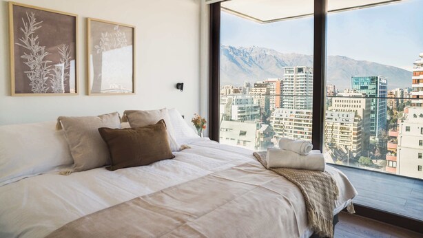 A bedroom with a large bed, two framed artworks, a window with a cityscape view, and a mountain in the background. - Prime 2BR Oasis in the Heart of Las Condes (Unknown)