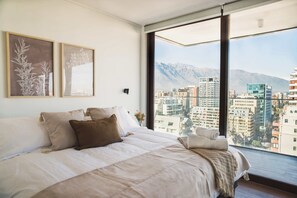 A bedroom with a large bed, two framed artworks, a window with a cityscape view, and a mountain in the background. - Prime 2BR Oasis in the Heart of Las Condes (Unknown)