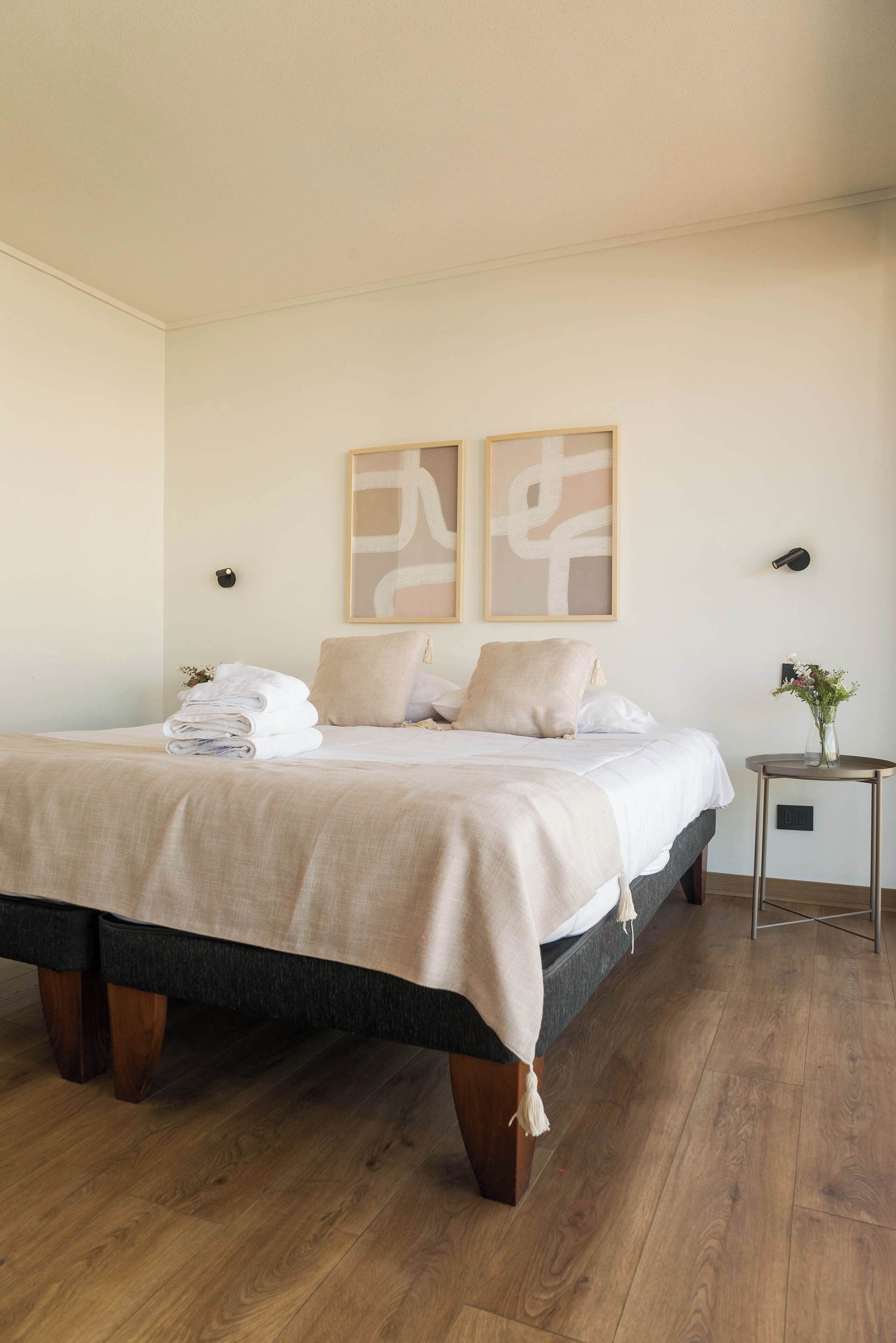 A neatly made bed with a beige blanket and pillows, a small bedside table with a vase of flowers, and abstract wall art above the bed.