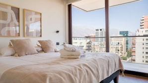 Een slaapkamer met een groot bed, twee ingelijste abstracte schilderijen en een uitzicht op een stadslandschap door een groot raam.