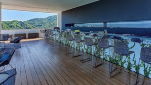 Um terraço amplo ao ar livre com piso de madeira, cadeiras de vime e um espaço de bar com vista para a cidade e as colinas.