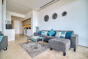 A modern living room with a grey sofa, a glass coffee table, and a grey rug. There is a kitchen in the background with wooden cabinets and a white countertop. - Modern Corner Condo with Epic Pool at SOHO (Puerto Vallarta)