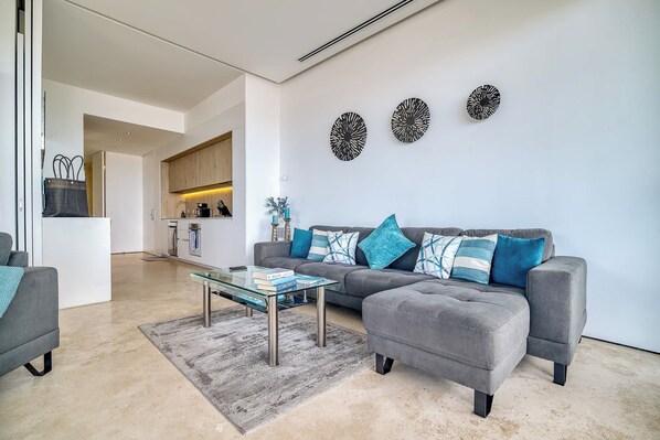 Una sala moderna con un sofá gris, una mesa de centro de vidrio y una alfombra gris. Se ve una cocina al fondo con armarios de madera y una encimera blanca.