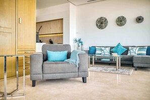 A modern living room with a grey armchair, a sofa with blue and white cushions, and a glass coffee table. There are decorative wall art pieces and a kitchen area in the background.