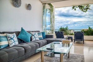 Un moderno salón con un sofá gris, cojines azules y blancos, una mesa de centro de vidrio y vistas a la ciudad y al mar a través de amplias ventanas.