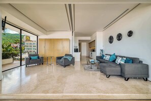 Amplia sala de estar con un sofá grande de color gris, un sillón gris más pequeño y una mesa de centro. Hay una puerta corrediza de vidrio que da a un balcón con vistas a un paisaje verde y edificios.