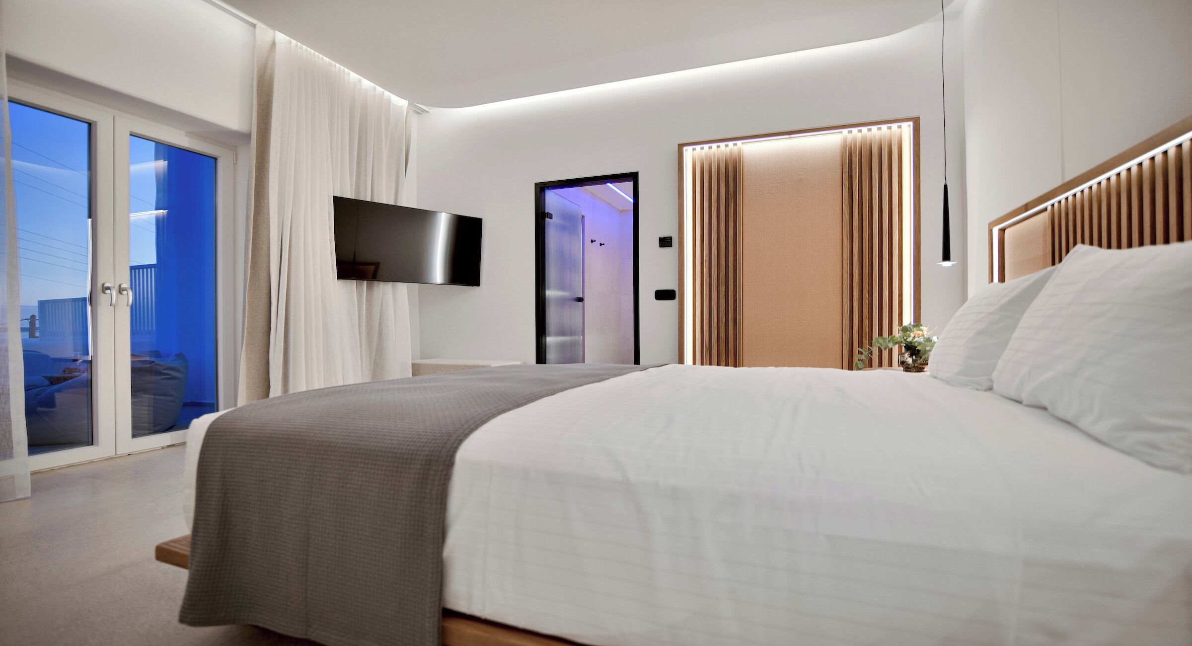 A modern hotel room with a large bed, a flat-screen TV, a sliding door to a balcony, and a shower area.