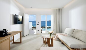 A modern hotel room with a large window offering a sea view, a flat-screen TV, a wooden table, a sofa, and a small round table with a vase.