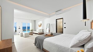 Una habitació d'hotel moderna amb un llit gran, una taula de nit amb un plat de petitdejuni, una zona d'estar amb un sofà i cadires, i una vista al mar a través de grans finestres.
