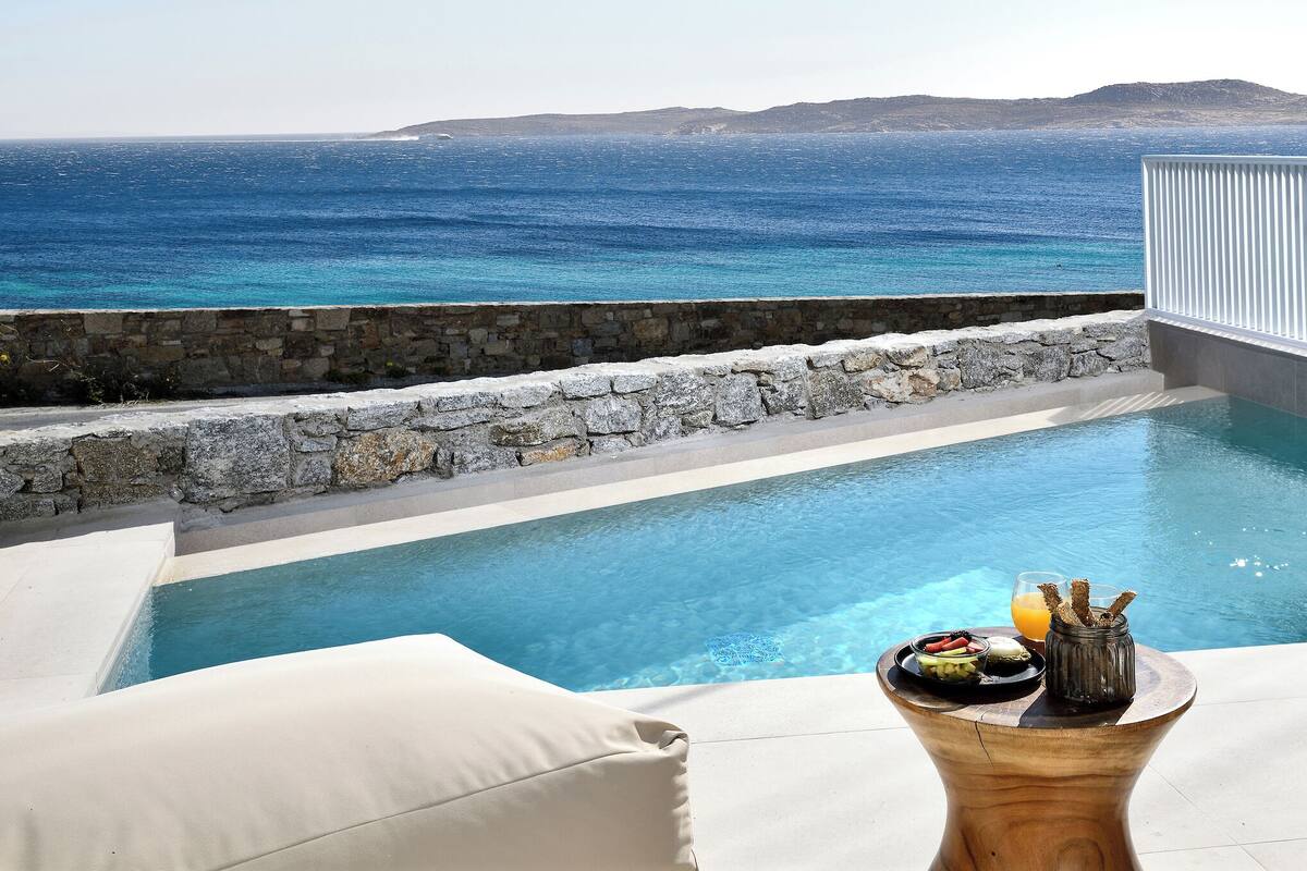 A pool with a stone wall and a view of the sea. There is a table with food and drinks.
