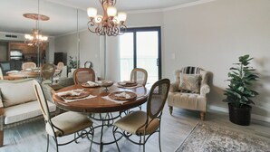 Un coin repas avec une table ronde, des chaises et un lustre. Un grand miroir orne un des murs, et une plante en pot se trouve dans un coin.