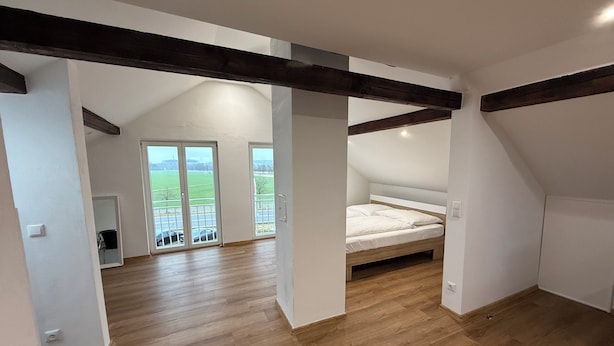 A bedroom with a sloped ceiling, wooden beams, a bed, and a balcony with a view of the countryside. - 1-bedroom apartment in Vilseck with WiFi, walking distance to base & food trucks (Vilseck)