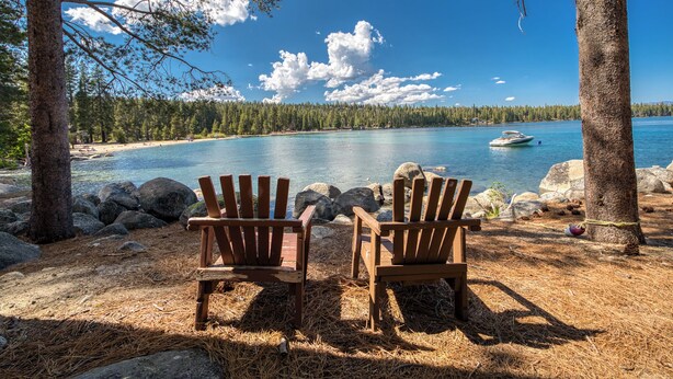 Two wooden Adirondack chairs facing a calm lake with a forested shoreline and a boat in the distance.