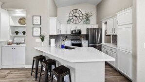 Une cuisine moderne avec des meubles blancs, un îlot central doté de tabourets, une grande horloge et un élément décoratif mural.